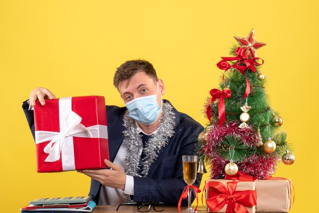 Front view of business man holding gift sitting at the table near xmas tree and presents on yellow