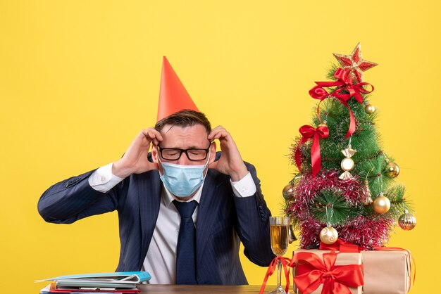Front view of business man holding his head sitting at the table near xmas tree and presents on yellow