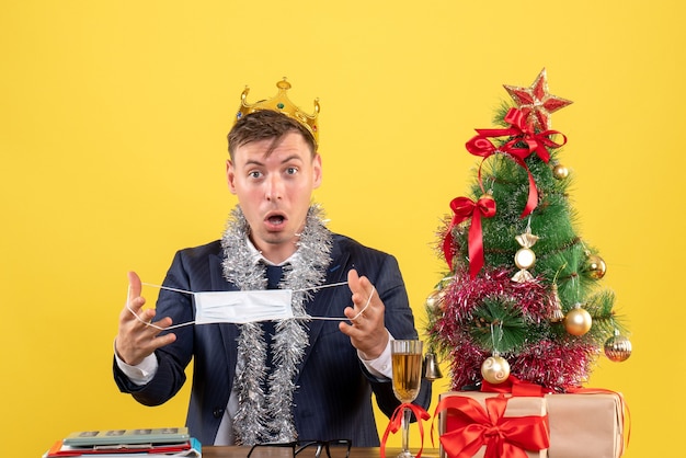 Front view of business man holding his mask sitting at the table near xmas tree and presents on yellow