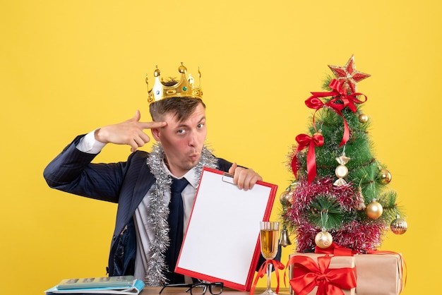 Vista frontale dell'uomo di affari che tiene la pistola del dito al suo tempiale che si siede al tavolo vicino all'albero di natale e presenta su colore giallo