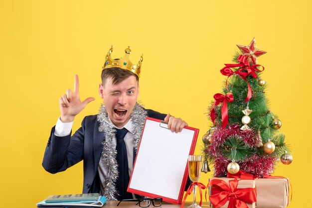 Front view of business man making finger gun sitting at the table near xmas tree and presents on yellow