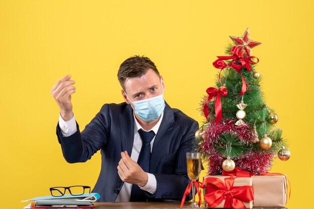 Front view of business man making money sign sitting at the table near xmas tree and presents on yellow