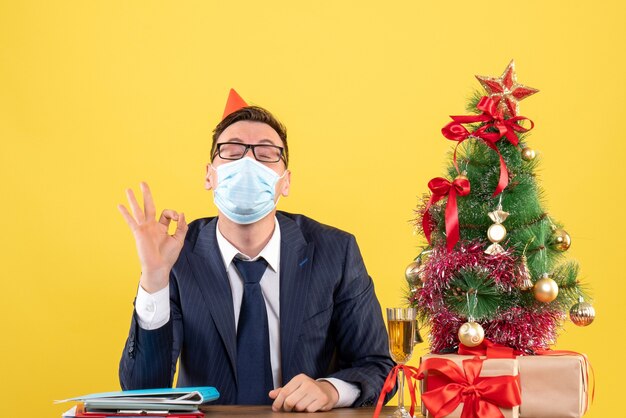 Front view of business man making ok sign sitting at the table near xmas tree and presents on yellow