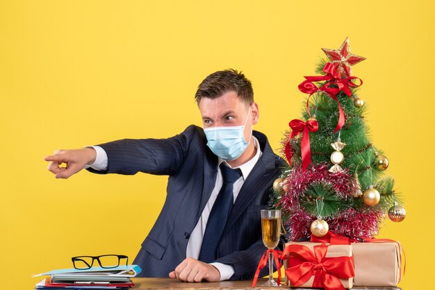 Front view of business man pointin at something sitting at the table near xmas tree and presents on yellow