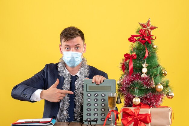 Front view of business man pointing at calculator sitting at the table near xmas tree and presents on yellow