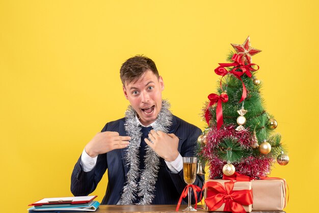 Front view of business man pointing at himself sitting at the table near xmas tree and presents on yellow
