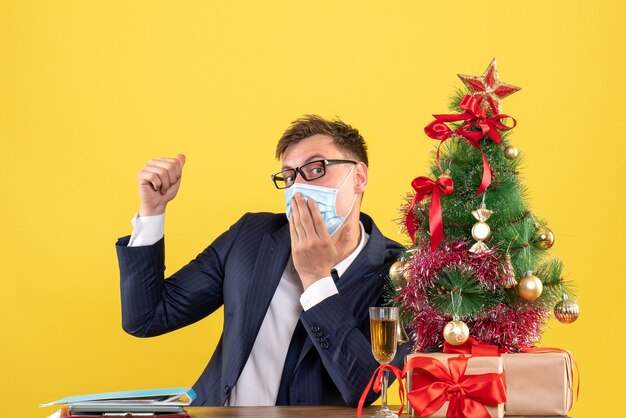 Front view of business man putting hand on his mouth sitting at the table near xmas tree and presents on yellow