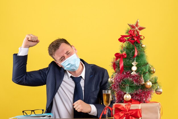 Front view of business man raising his hand sitting at the table near xmas tree and presents on yellow