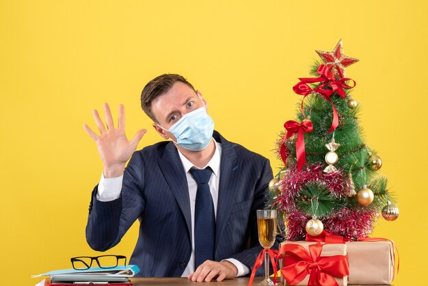 Front view of business man saying hello sitting at the table near xmas tree and presents on yellow