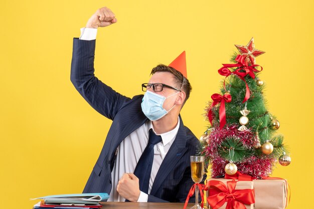 Front view of business man sitting at the table near xmas tree and presents on yellow.