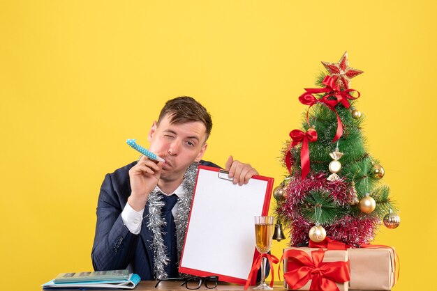 Front view of business man using noisemaker sitting at the table near xmas tree and presents on yellow