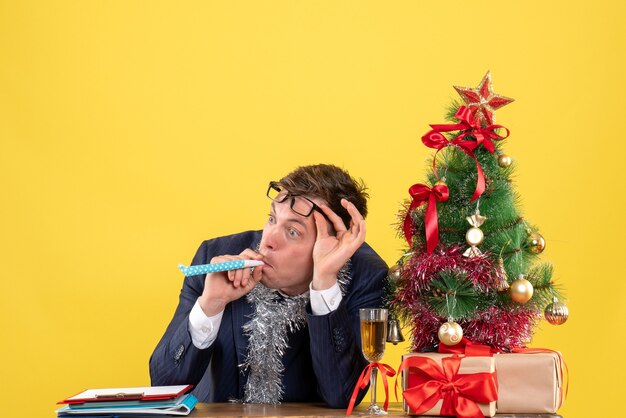 Front view of business man using noisemaker sitting at the table near xmas tree and presents on yellow