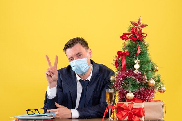 Front view of business man with blinked eye making victory sign sitting at the table near xmas tree and presents on yellow