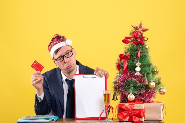 Front view of business man with blinked eye sitting at the table near xmas tree and presents on yellow