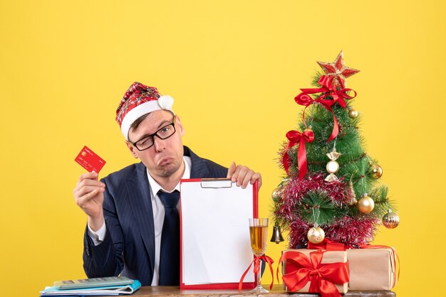 Front view of business man with blinked eye sitting at the table near xmas tree and presents on yellow