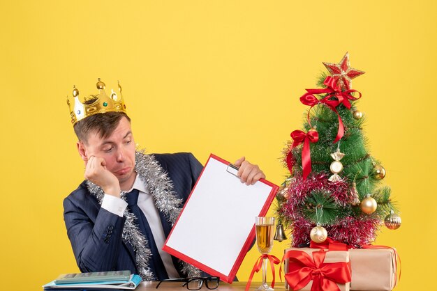 Front view of business man with crown checking paper sitting at the table near xmas tree and presents on yellow