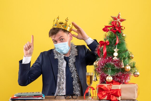 Front view of business man with crown pointing at high sitting at the table near xmas tree and presents on yellow