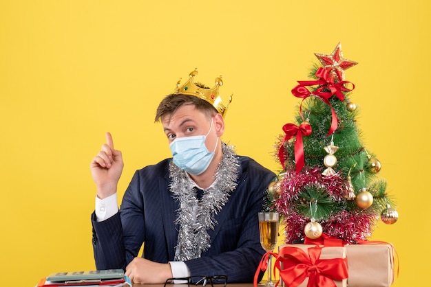 Vista frontale dell'uomo di affari con la corona che si siede al tavolo vicino all'albero di natale e regali su giallo