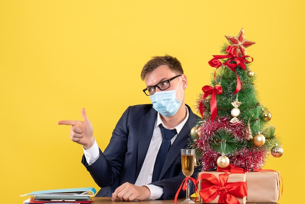 Front view of business man with eyeglasses making finger gun sitting at the table near xmas tree and presents on yellow