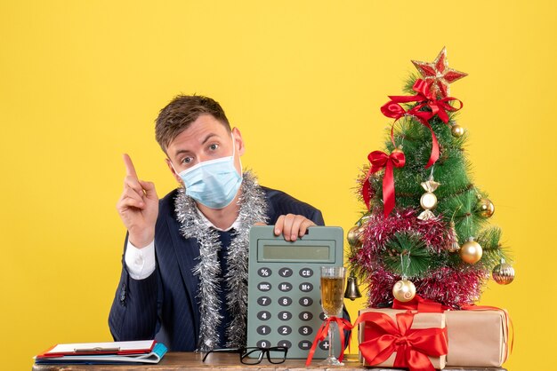 Front view of business man with mask pointing at calculator sitting at the table near xmas tree and presents on yellow