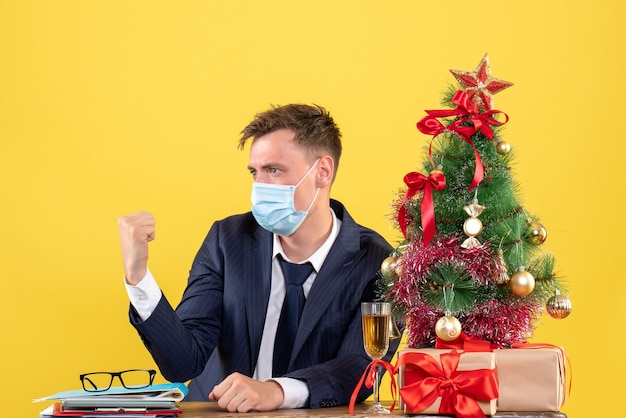 Вид спереди делового человека с медицинской маской, сидящего за столом возле рождественской елки и подарков на желтом