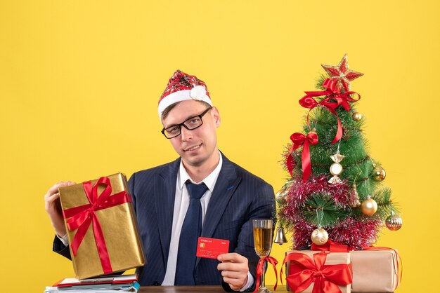 Front view of business man with santa hat holding card and gift sitting at the table near xmas tree and presents on yellow