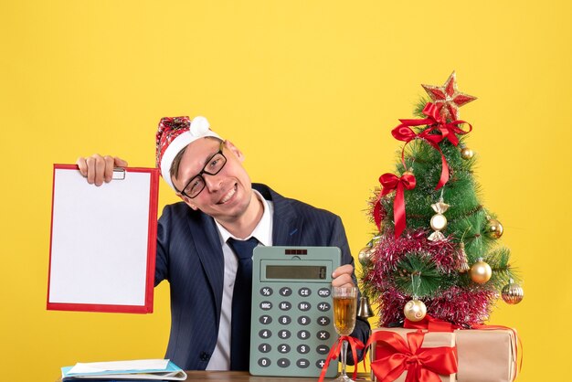 Front view of business man with santa hat holding clipboard and calculator sitting at the table near xmas tree and presents on yellow