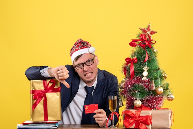 산타 모자는 크리스마스 트리 근처 테이블에 앉아 엄지 손가락을 만드는 비즈니스 남자의 전면보기와 노란색 선물