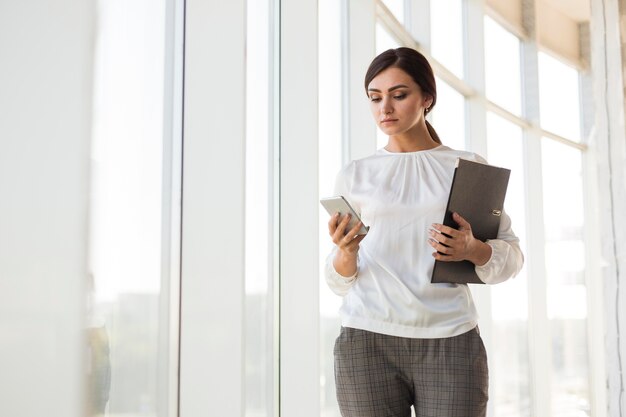 Front view of businesswoman holding binder and looking at smartphone