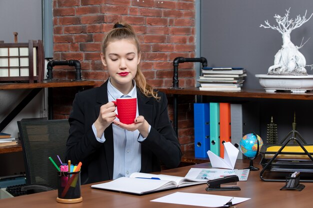 Front view of busy young female sitting at a table and holding a red cup in the office