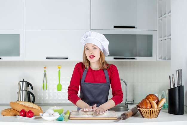 Вид спереди занятая молодая женщина в поварской шляпе и фартуке замешивает тесто на кухне