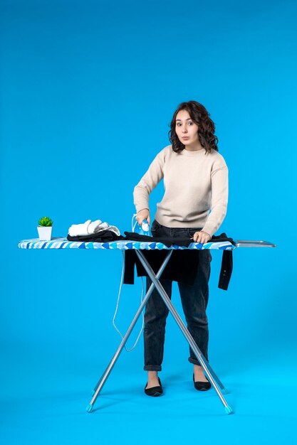 Front view of busy young woman standing behind the board and ironing the cloth on blue wave surface