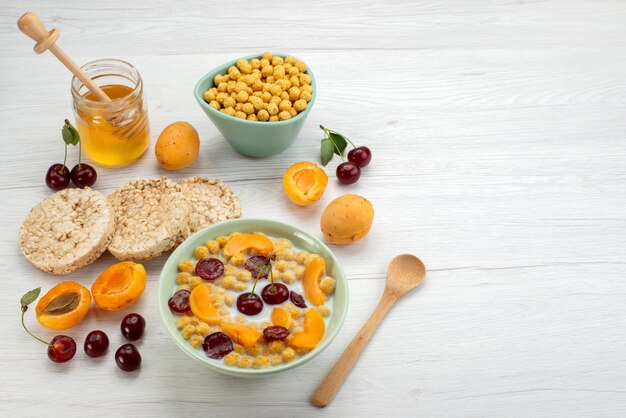 Вид спереди хлопья с молоком внутри синей тарелки с крекерами, фруктами и медом на белом столе пить молоко, молочные сливки, завтрак