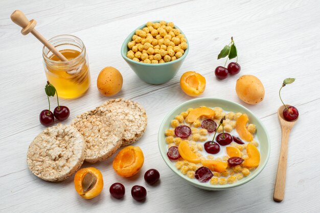 Вид спереди хлопья с молоком внутри тарелки с крекерами, фруктами и медом на белом столе, пейте молочные сливки