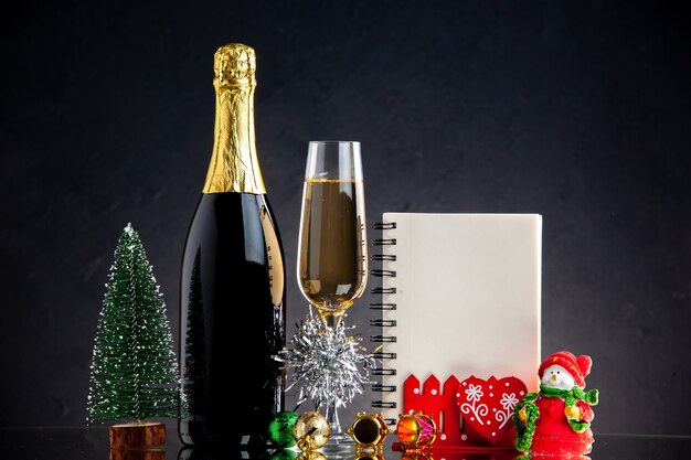 Front view champagne glass bottle xmas ornaments notebook mini xmas tree on dark surface