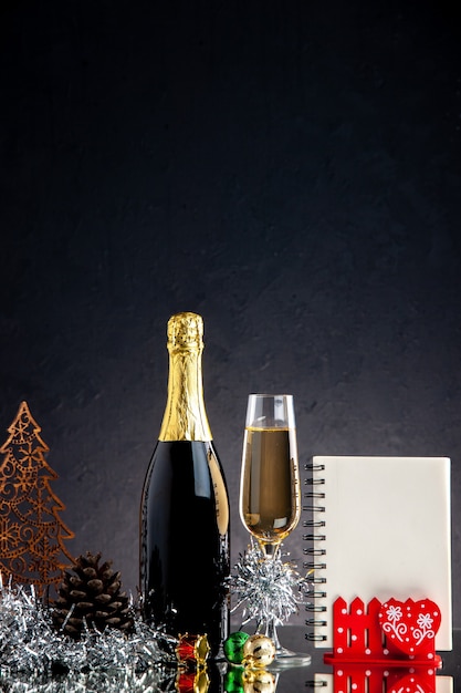 Стеклянная бутылка шампанского, вид спереди, рождественские украшения, блокнот на темной поверхности