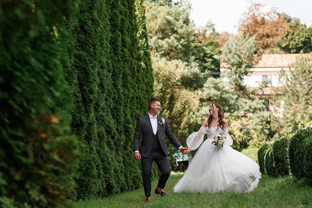 緑の中をお散歩する陽気な愛情のある花嫁の正面図ウェディングドレスを着て花の花束を持っている美しい花嫁お互いを見ている夫婦
