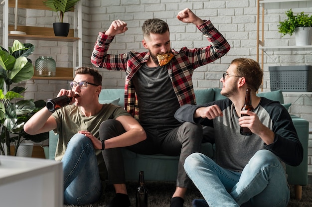 Front view of cheerful male friends having pizza and watching sports on tv