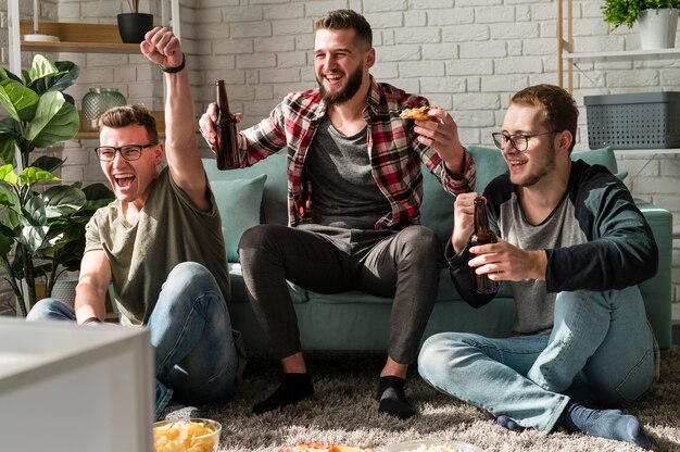 Вид спереди веселых друзей-мужчин, которые едят пиццу с пивом и смотрят спорт по телевизору
