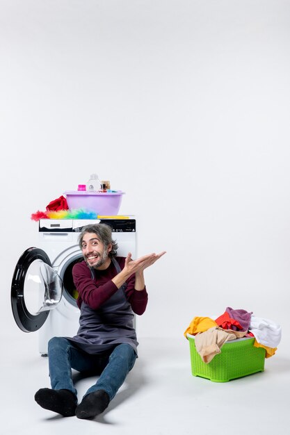Front view cheerful male housekeeper sitting in front of washer laundry basket on white background