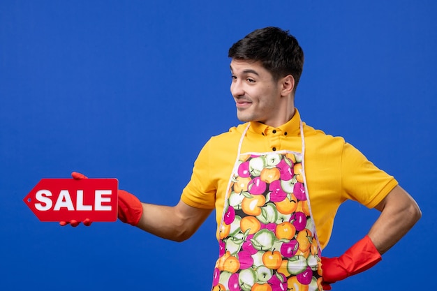 Front view cheerful male housekeeper in yellow t-shirt holding sale sign putting hand on a waist on blue space