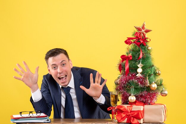 Вид спереди веселого человека, открывающего руки, сидящего за столом возле рождественской елки и подарков на желтом