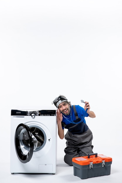 Вид спереди веселый ремонтник, держащий стетоскоп, сидя возле стиральной машины на белом пространстве