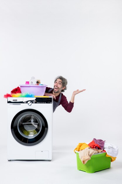 Front view cheerful young man in apron sitting behind washer laundry basket on white background