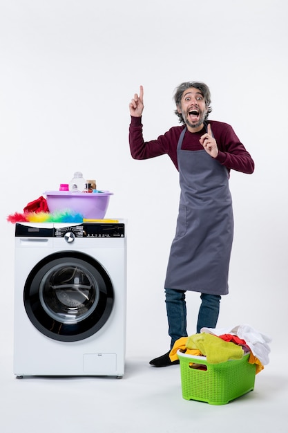 Front view cheery housekeeper man pointing at ceiling standing near washer laundry basket on white background