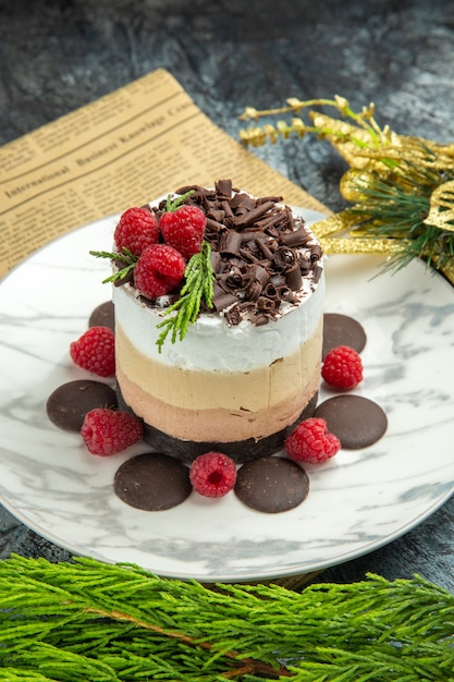 Front view cheesecake with chocolate and raspberries on white oval plate on newspaper xmas ornaments on grey background