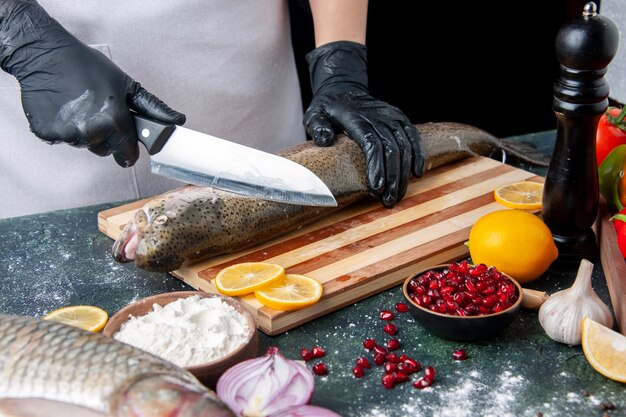 Вид спереди шеф-повар, измельчающий сырую рыбу на деревянной доске, мельница для перца, миска для муки, семена граната в миске на кухонном столе