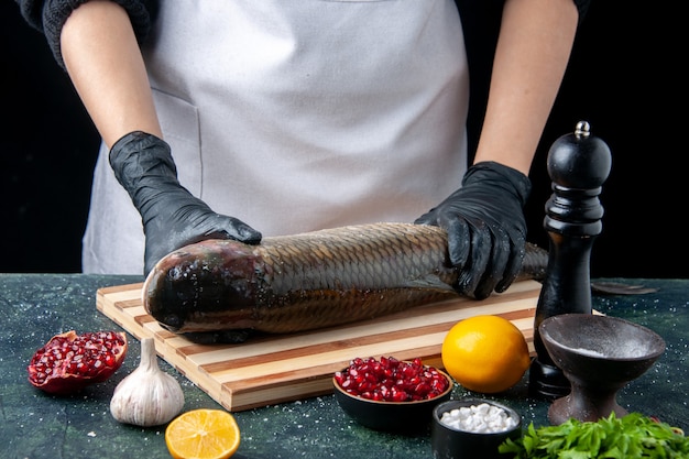 Free photo front view chef in holding raw fish on chopping board pepper grinder pomegranate seeds in bowl on table