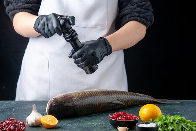 Free photo front view chef in white apron sprinkled pepper with pepper grinder onto fresh fish pomegranate seeds in bowl on table