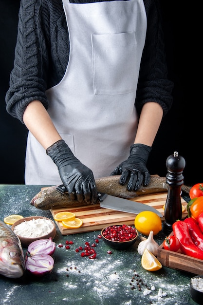 Бесплатное фото Шеф-повар, вид спереди в черных перчатках, рубит сырую рыбу на деревянной доске, мельница для перца, миска для муки, семена граната в миске на кухонном столе